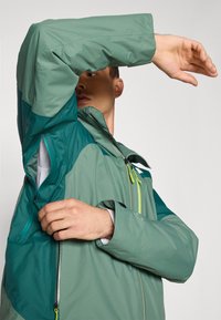 Veste verte et turquoise avec une poche zippée, en tissu texturé et avec des manches ajustables. Présente un col haut et des contrastes de couleur sur les épaules.