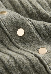 Grüner strukturierter Stoff mit geripptem Muster, versehen mit großen goldenen Knöpfen, die einen Kontrast zum Material bilden. Nahaufnahme hebt die Details hervor.