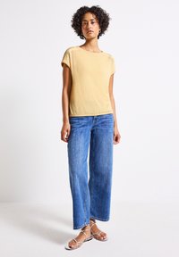 Ljust gult texturerat kortärmat plagg i kombination med löst sittande, medblå denimjeans och platta silverfärgade sandaler med remmar, uppvisat mot en enfärgad bakgrund.