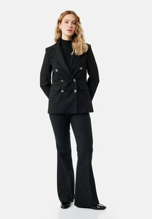 Femme aux cheveux blonds ondulés portant un blazer noir croisé, un pantalon noir évasé et des chaussures noires, debout les mains dans le dos.