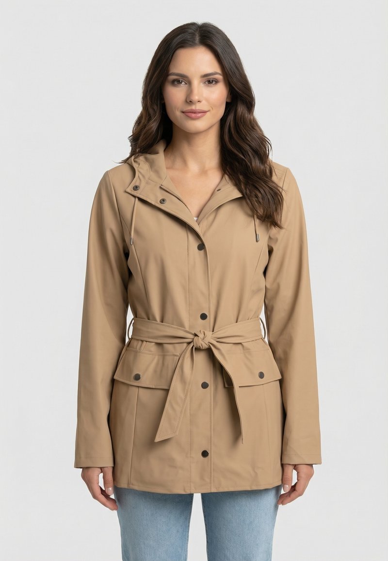 Donna con capelli lunghi e scuri che indossa una giacca beige con cintura, bottoni frontali e tasche a pattina, in piedi davanti a uno sfondo chiaro uniforme.