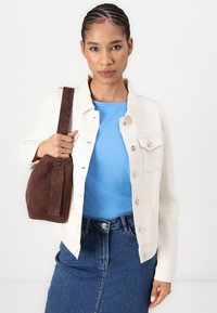 Hvit dongeri jakke med metallknapper, parret med en blå ribbet topp og en brun semsket håndveske. Modellen har på seg et dongeri skjørt.