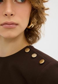 Bruine stoffen top met ronde, gestructureerde gouden knopen op de schouders. Glad textuur, minimalistisch ontwerp met goed zichtbare gelaatstrekken.