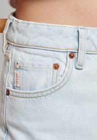 Pantalones cortos de mezclilla azul claro de talle alto con el dobladillo deshilachado, cierre en el botón superior y un pequeño bolsillo delantero. Presenta costuras amarillas y una etiqueta de la marca.