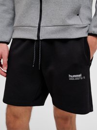 Homme portant un short de sport noir Hummel avec des poches latérales et une veste zippée grise, les mains dans les poches, debout devant un fond uni.