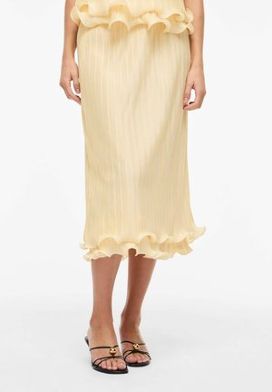 Lys gul plisseret midi-nederdel med flæset kant, båret med sorte sandaler med stropper og guldpynt på hvid baggrund.