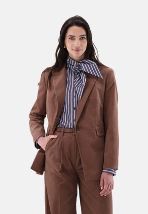 Femme en costume marron avec ceinture assortie porte une chemise bleue rayée et un grand nœud noué au cou, debout avec une main dans la poche.