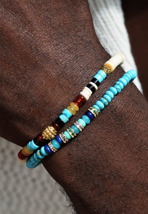 Drei Armbänder an einem Handgelenk: türkisblaue Perlen, goldene Akzente, Elemente mit schwarz-weißen Mustern, gemischte Materialien, unterschiedliche Größen und Formen.