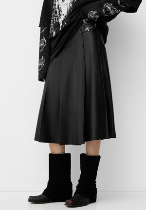 Femme portant une jupe midi plissée noire avec des détails de boucle latérale, un t-shirt graphique noir à manches longues, et des bottes noires tombantes.