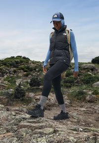 Gilet trapuntato nero sopra una camicia a maniche lunghe azzurro chiaro, leggings neri, calzini grigi e scarpe da trekking nere; circondato da un terreno roccioso e vegetazione.
