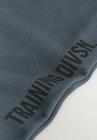 Tissu bleu avec texte imprimé en noir « TRAINING DIVISI », partiellement visible sur une surface courbée.