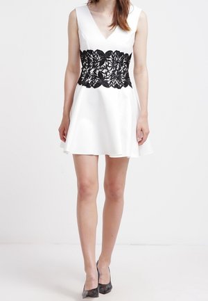 Weißes Kleid mit V-Ausschnitt und ausgestelltem Rock. Schwarzer Spitzenbund mit floralen Mustern. Getragen mit schwarzen Absätzen. Glatte Stofftextur.