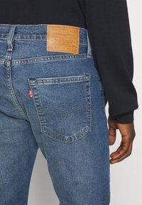 Vue de dos d'une personne portant un jean Levi's bleu avec une étiquette en cuir et une étiquette rouge Levi's sur la poche, vêtue d'un haut noir à manches longues.