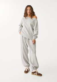 Hellgrauer Sweatshirt mit einem schulterfreien Design, lockeren Ärmelbündchen und passenden, lockeren grauen Jogginghosen. Braune Sneaker vervollständigen das Outfit.