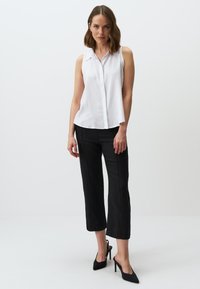 Blusa bianca senza maniche con bottoni frontali e colletto, abbinata a pantaloni neri cropped e scarpe con tacco nere.