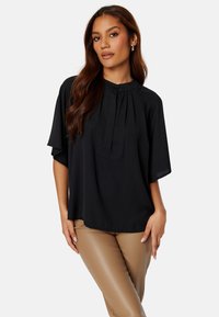 Blusa nera a maniche corte con colletto, pieghe e vestibilità rilassata. Tessuto morbido e materiale leggero. Abbinata a pantaloni beige aderenti.