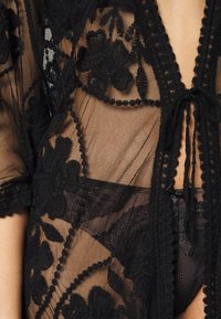 Czarny koronkowy kimono z kwiecistym haftem, przejrzysty materiał, zawiązywany w talii i z falbaniastymi krawędziami. Posiada delikatną fakturę i złożone detale.