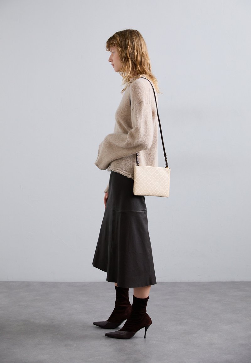 Beige gestrickter Pullover mit weiten Ärmeln, schwarze Leder-Midiröcke und dunkelbraune Ankle-Boots mit spitzen Zehen. Weiße Umhängetasche mit einem Muster.