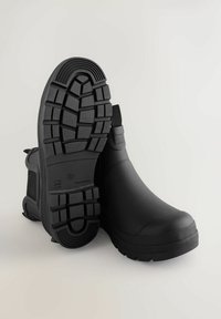 Schwarze wasserfeste Knöchelstiefel verfügen über eine strukturierte Gummisohle mit tiefem Profil, elastische Seiteneinsätze und ein glattes Obermaterial.
