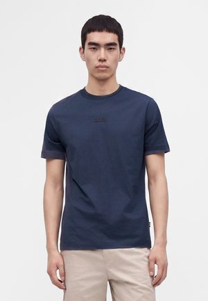 Young man with short black hair wearing a navy blue BOSS t-shirt and beige pants standing against a plain white background.