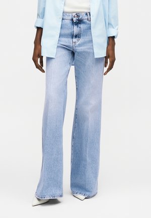 Ανοιχτό μπλε τζιν με ψηλή μέση και φαρδιά μπατζάκια, φορεμένο με λευκά μυτερά παπούτσια, λευκό μπλουζάκι και ανοιχτό μπλε πουκάμισο με γυρισμένα μανίκια.