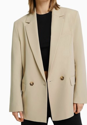 Mujer con blazer beige de doble botonadura con botones de carey sobre top negro, recortada a la mitad del muslo, manos relajadas a los costados.