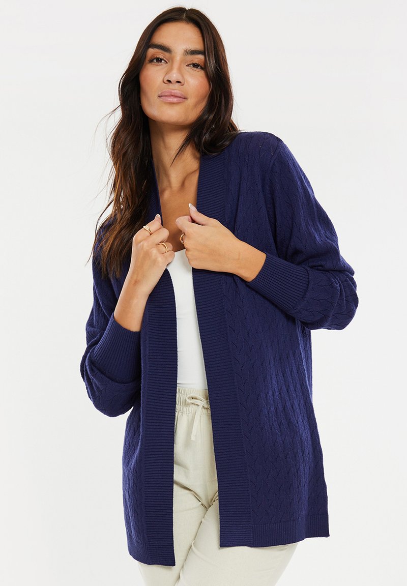 Cardigan azul marino hecho de un tejido de punto texturizado con un patrón de trenzas, de frente abierto y con puños de canalé. Lleva puesto un top blanco.