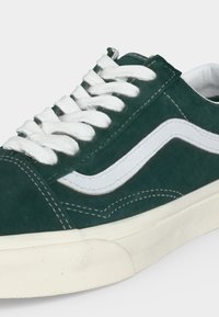 Ciemnozielony zamszowy sneaker z białym paskiem po boku, białymi sznurowadłami, widocznym przeszyciem i kremową gumową podeszwą.