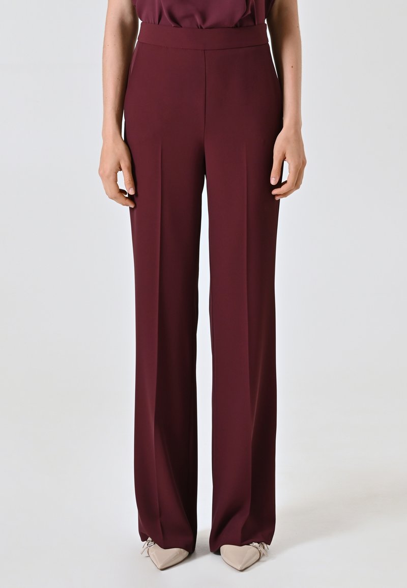 Bordeauxfarbene weit geschnittene Hosen mit glatter Textur, hoher Taille und dezenten Falten vorne, kombiniert mit beigen, spitz zulaufenden Ballerinas.