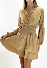 Robe en satin beige avec manches longues, taille froncée, décolleté en V, et jupe évasée ornée de volants superposés. Texture lisse et brillante.