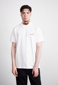 T-shirt bianca in cotone con maniche corte. Presenta un piccolo logo Carhartt rosso e giallo sul lato sinistro del petto. Design semplice, vestibilità comoda.