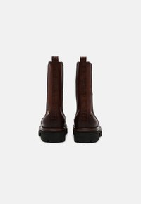 Une paire de bottes en cuir marron foncé, hautes, avec une bande texturée à l'arrière, des tirettes en boucle, des semelles noires épaisses et de petits embellissements métalliques près des talons.