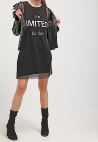 Svart nätklänning med texten "Jag är en BEGRÄNSAD upplaga", lagd under en svart läderjacka. Snygg, figurnära form med knälång fåll.