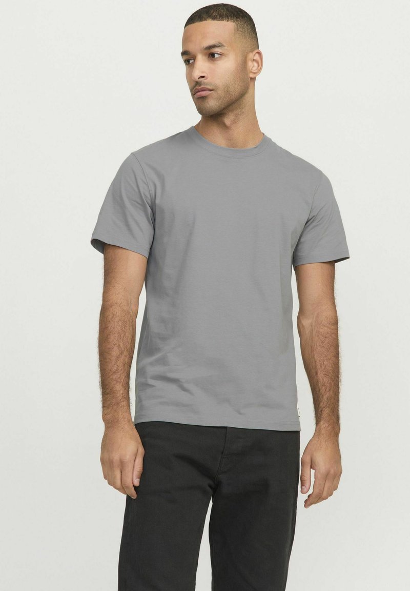 Camiseta de algodón gris de manga corta con cuello redondo. El modelo la lleva con pantalones negros, mostrando un ajuste relajado.
