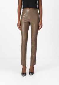 Pantaloni in finta pelle marrone con un design aderente, caratterizzati da una vita alta, gambe slim e dettagli di cucitura minimi. Indossati con tacchi alti neri.