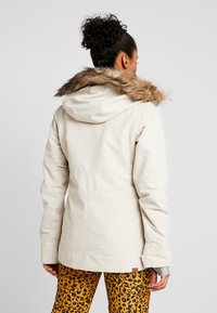 Helle beige Jacke mit einer mit Fell gefütterten Kapuze, mit tailliertem Schnitt, flachen Nähten und glatter Textur. Kombiniert mit Hose im Leopardenmuster.