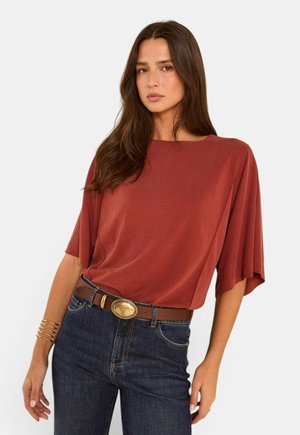 Femme aux longs cheveux bruns portant un haut ample couleur rouille, un jean bleu foncé, une ceinture marron avec boucle dorée et des bracelets dorés au poignet gauche.