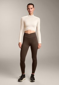 Beige pitkähihaine crop-top, jossa on epäsymmetrinen helma, yhdistettynä korkeavyötäröisiin ruskeisiin legginsseihin, joissa on V-muotoinen vyötärö ja sileä pinta.