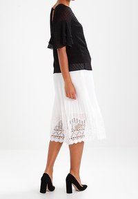 Blusa nera a pois con maniche a volant, abbinata a una gonna bianca plissettata con bordo in pizzo, completata da scarpe con tacco a blocco nere.