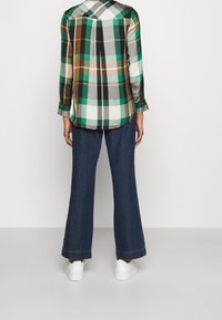 Camisa de cuadros con colores verde, marrón y gris, combinada con jeans de mezclilla azul de pierna ancha y zapatillas blancas, vista desde la parte de atrás.
