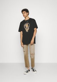 T-shirt noir surdimensionné avec un graphique circulaire au centre. Porté avec un jean déchiré marron clair et des baskets noir et blanc.