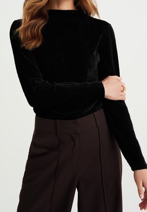 Sort langærmet top med tekstureret, ribbet design; kombineret med højtaljede brune bukser med skarpe presninger.