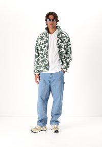 Veste en polaire à motifs verts et blancs, t-shirt blanc, jeans bleu clair et baskets beiges. Porté avec des lunettes de soleil réfléchissantes.