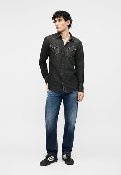 Jeune homme debout les mains dans les poches, portant une chemise en jean noire, un jean bleu et des baskets noires à rayures blanches.