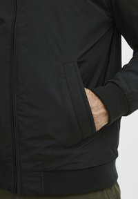 Črn bomberski jopič z gladko, sijočo teksturo, rebrastimi manšetami in robom ter stranskimi žepi. Ima zadrgo spredaj za zapiranje.