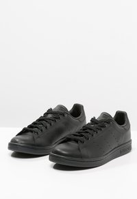 Sorte læder sneakers med runde tæer, flade snørebånd, perforerede detaljer og teksturerede gummisåler. Branding på hælen. Simpelt design.