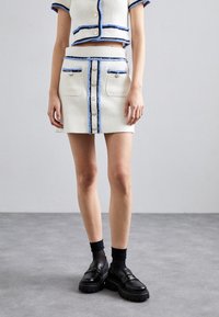 Witte tweed twee-delige outfit bestaande uit een cropped jack met blauwe randen en een bijpassende rok met knopen aan de voorkant en zakken. Zwarte chunky loafers gedragen.