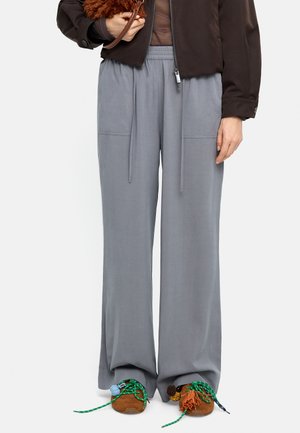 Osoba nosząca luźne szare spodnie i brązowe buty z kolorowymi zielonymi i pomarańczowymi sznurowadłami, trzymająca brązową puszystą torbę.