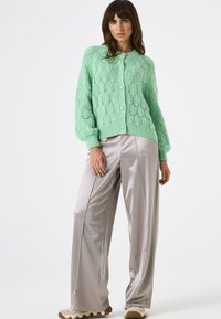 Hellgrüner, gestrickter Cardigan mit einem strukturierten Rautenmuster, Knopfverschluss und Puffärmel, kombiniert mit schimmernden silbernen weit geschnittenen Hosen.