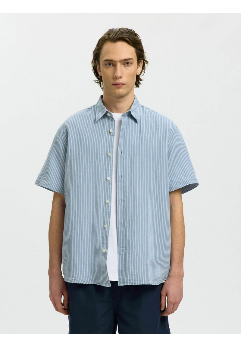 Jeune homme aux cheveux mi-longs portant une chemise à manches courtes rayée bleu clair et blanc par-dessus un t-shirt blanc et un pantalon sombre, debout devant un fond blanc.